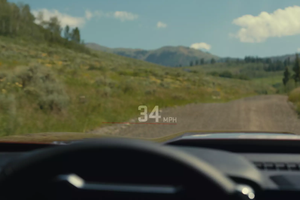 View from a GMC Canyon's head-up display showing '34 MPH' over a dirt road through green, hilly landscape.