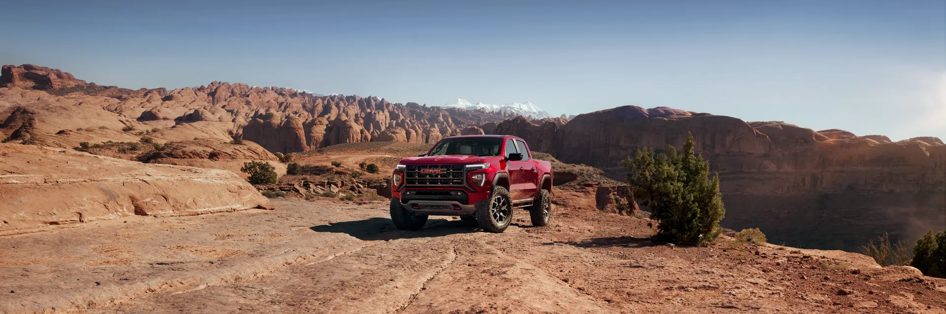 Red GMC pickup truck parked on rocky desert terrain with red sandstone formations and snowy mountains under a clear sky.
